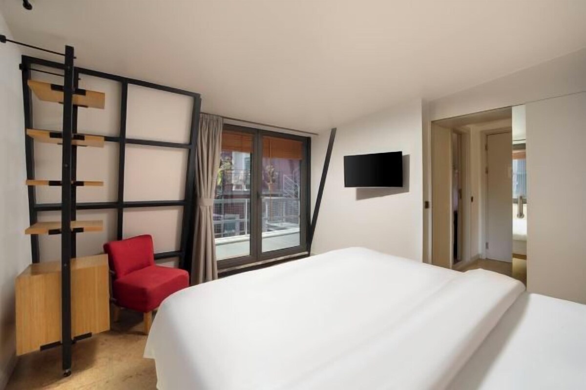 A hotel room with a bed, a red chair, a TV, and a window with a view.