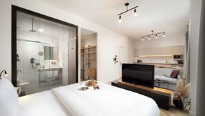 Une chambre d’hôtel moderne dotée d’un grand lit, d’une télévision, d’un petit coin cuisine et d’une salle de bain séparée par une paroi en verre.