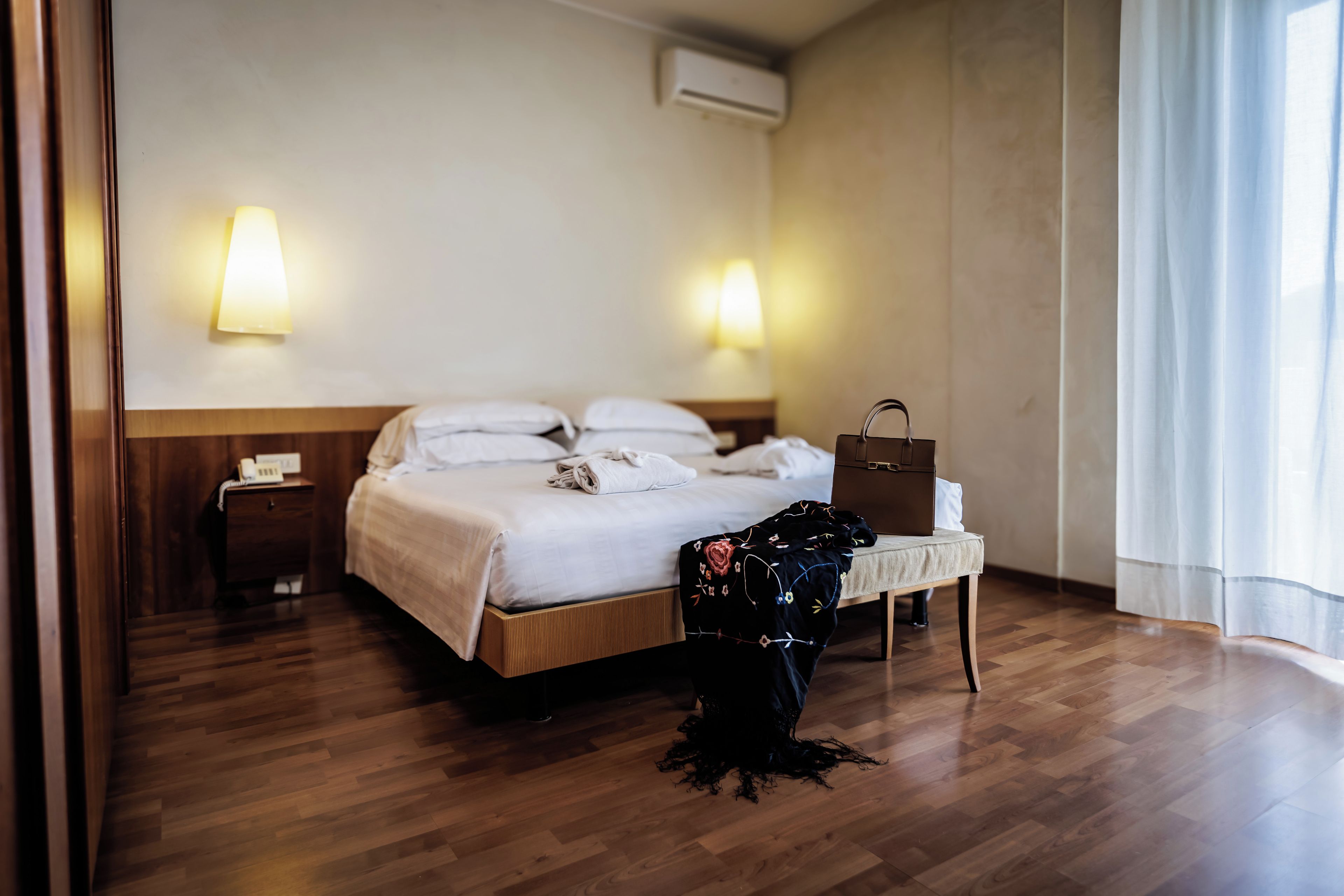 Habitación de hotel con piso de madera, cama con ropa blanca, mesita de noche con lámpara, maleta y ventana con cortinas.