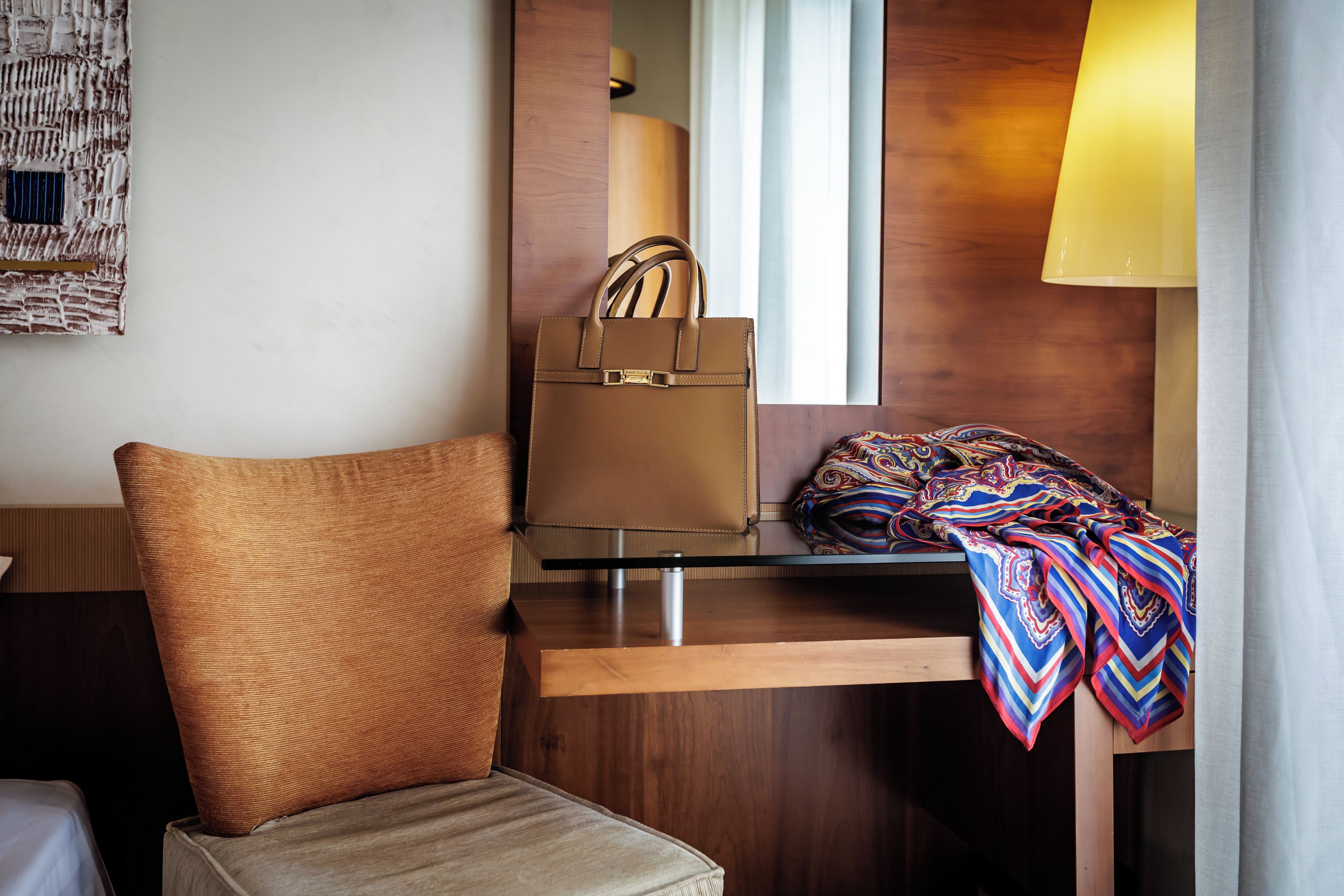 A hotel room with a brown leather chair, a wooden desk, a mirror, a lamp, and a colorful blanket.