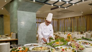 Ein Koch bereitet Essen in einer hellen, modernen Küche zu, auf einem Buffet sind verschiedene Speisen angerichtet.