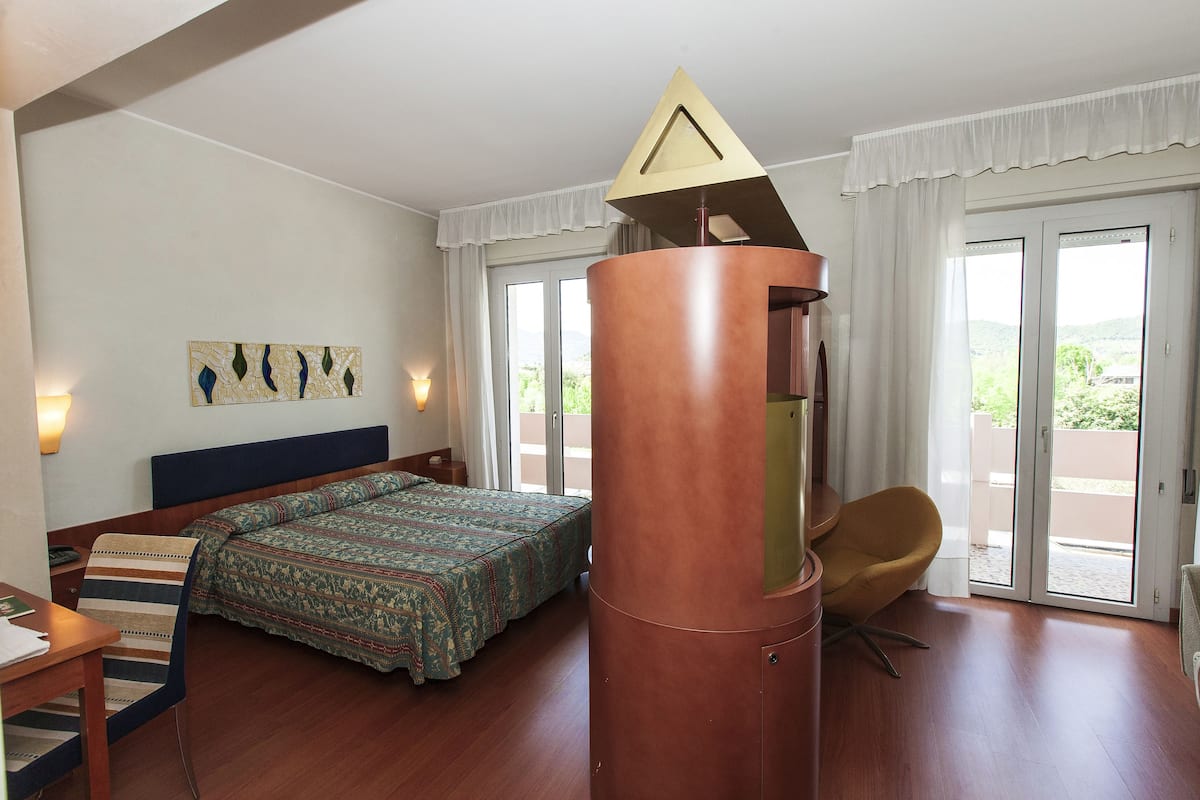 Ein Hotelzimmer mit Bett, Stuhl, Schreibtisch, Lampe, einem Gemälde und Blick ins Freie.