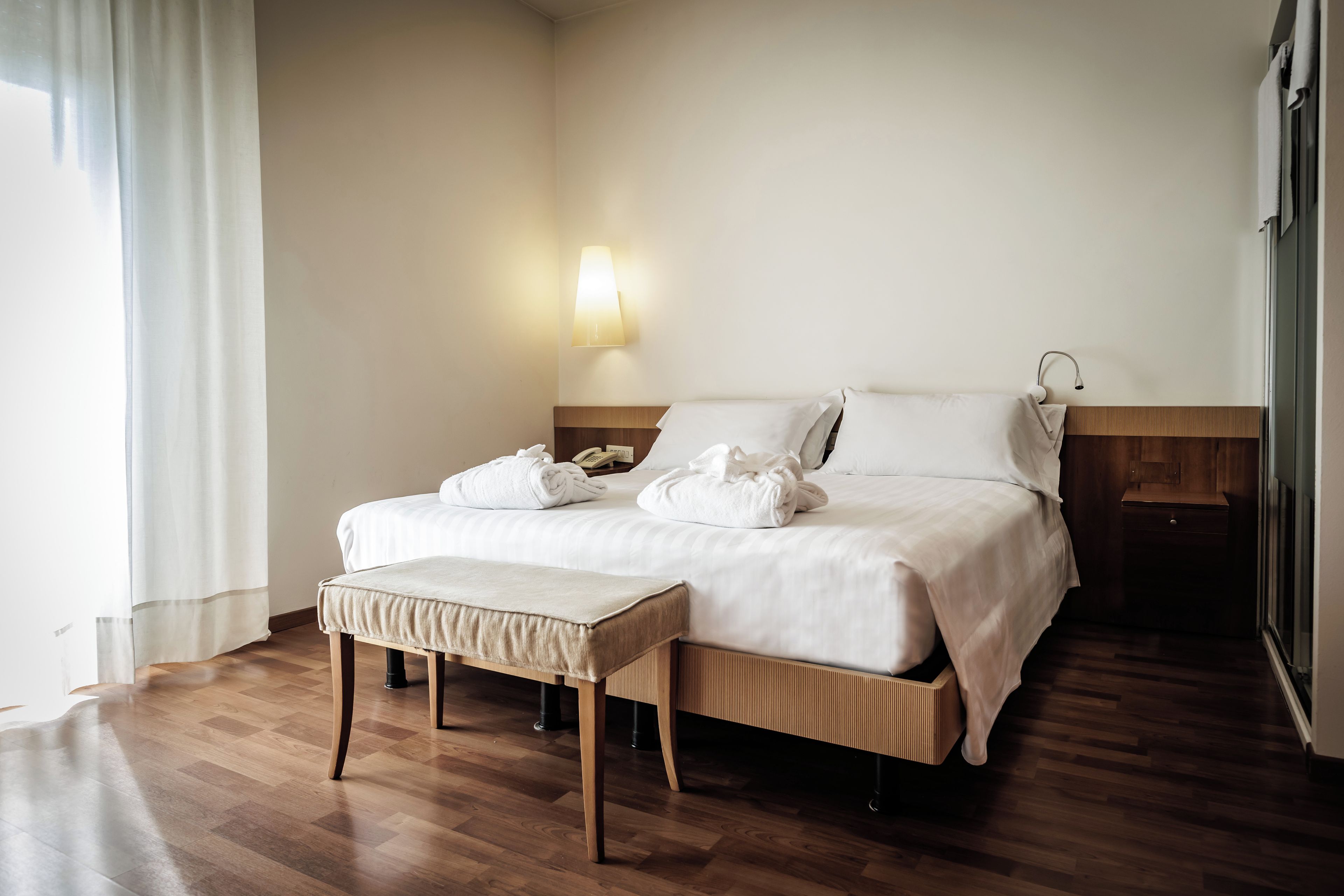 Quarto de hotel com cama, mesa de cabeceira, luminária e janela com cortinas.