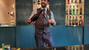 Eine Bar mit einer Theke, an der ein Barkeeper eine Flasche und ein Glas hält. Im Hintergrund sind Regale mit verschiedenen Spirituosen zu sehen.
