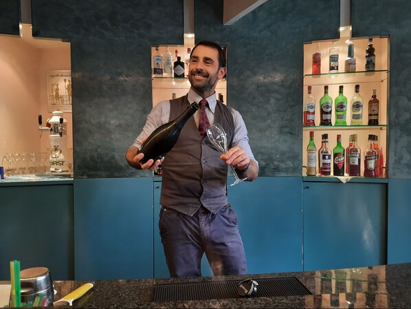 Eine Bar mit einer Theke, an der ein Barkeeper eine Flasche und ein Glas hält. Im Hintergrund sind Regale mit verschiedenen Spirituosen zu sehen.