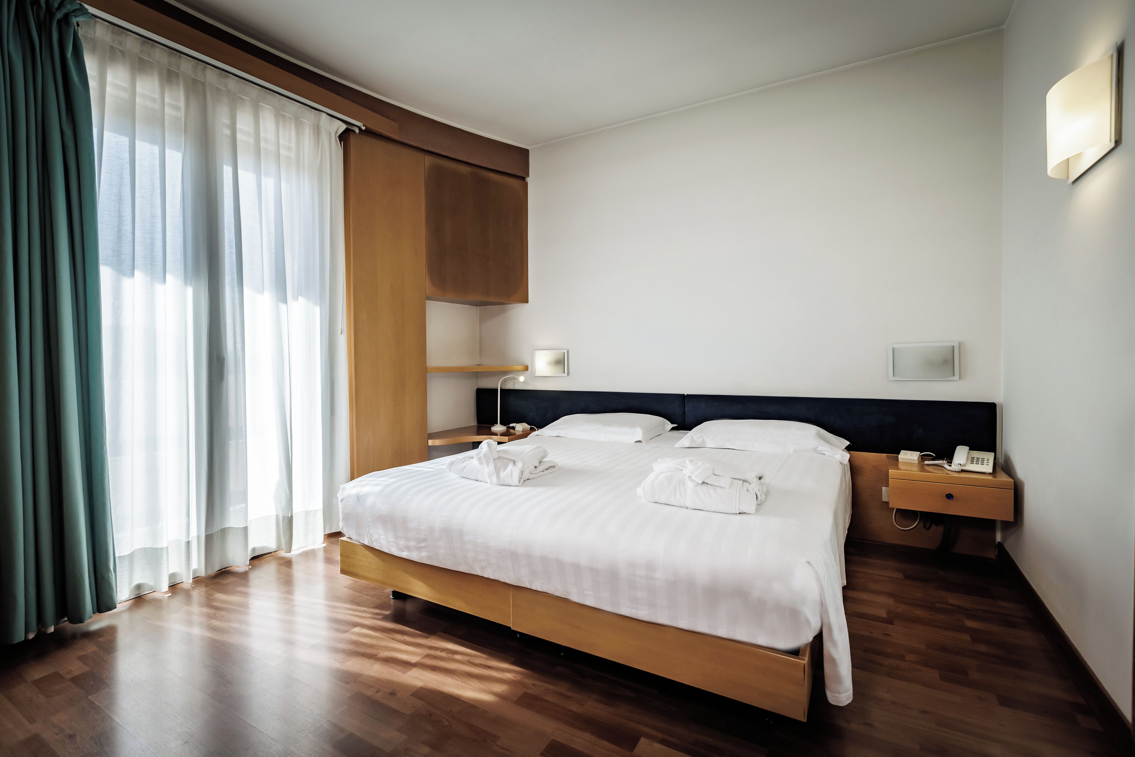 A hotel room with a large bed, wooden headboard, bedside tables with lamps, and a window with curtains.