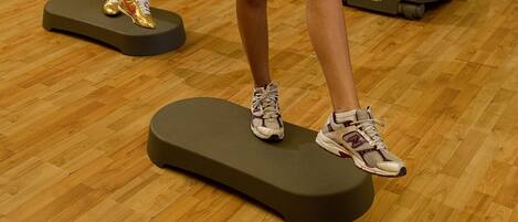 Dos personas ejercitándose en plataformas elevadas en una habitación con suelo de madera y equipo de gimnasio al fondo.