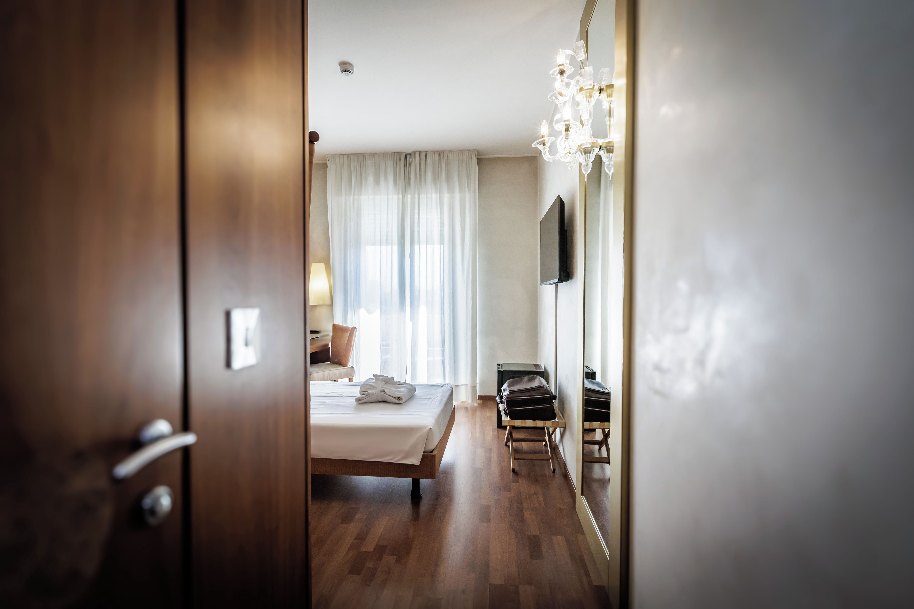 A hotel room with a bed, a bedside table, a lamp, a mirror, and a chandelier.