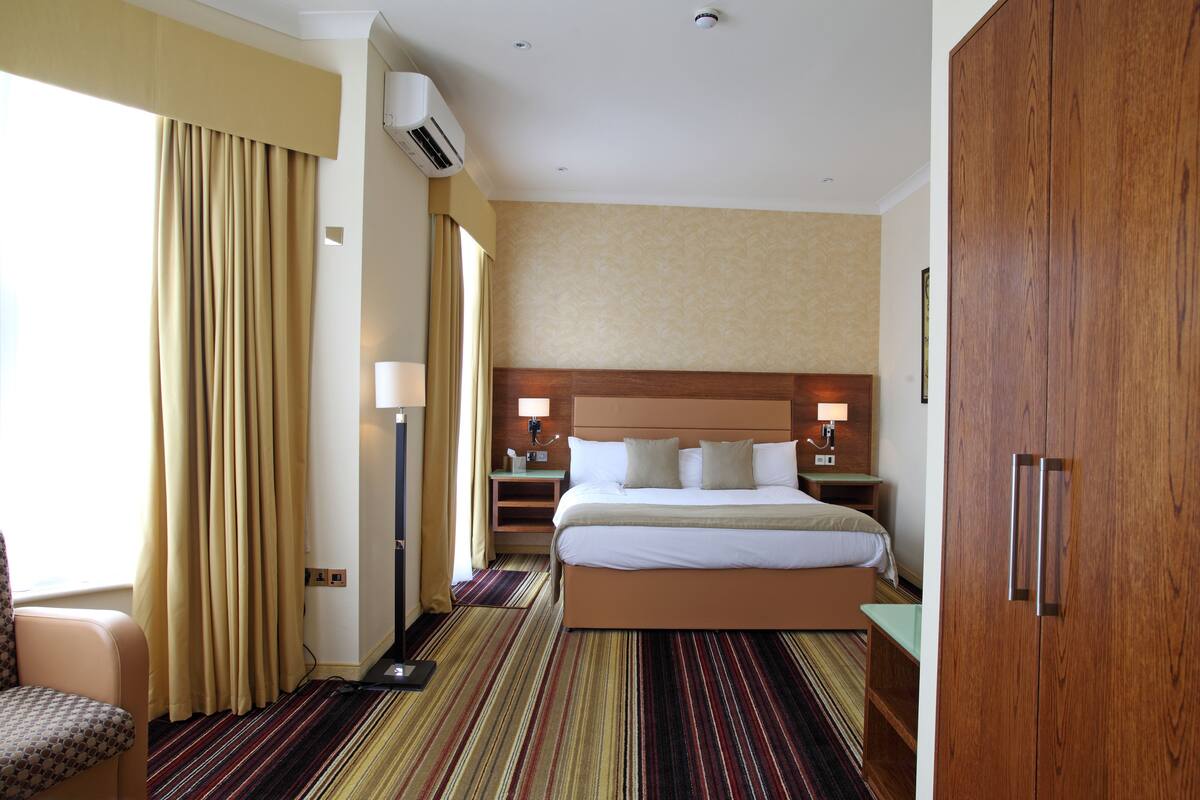A hotel room with a large bed, a wooden wardrobe, a desk with a lamp, and a chair.