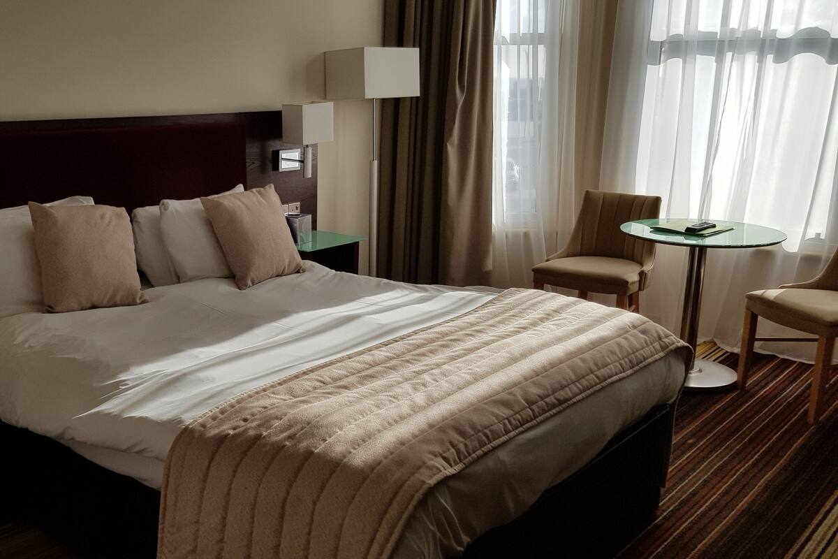A hotel room with a large bed, two bedside tables, a chair, a small table, and a window with curtains.