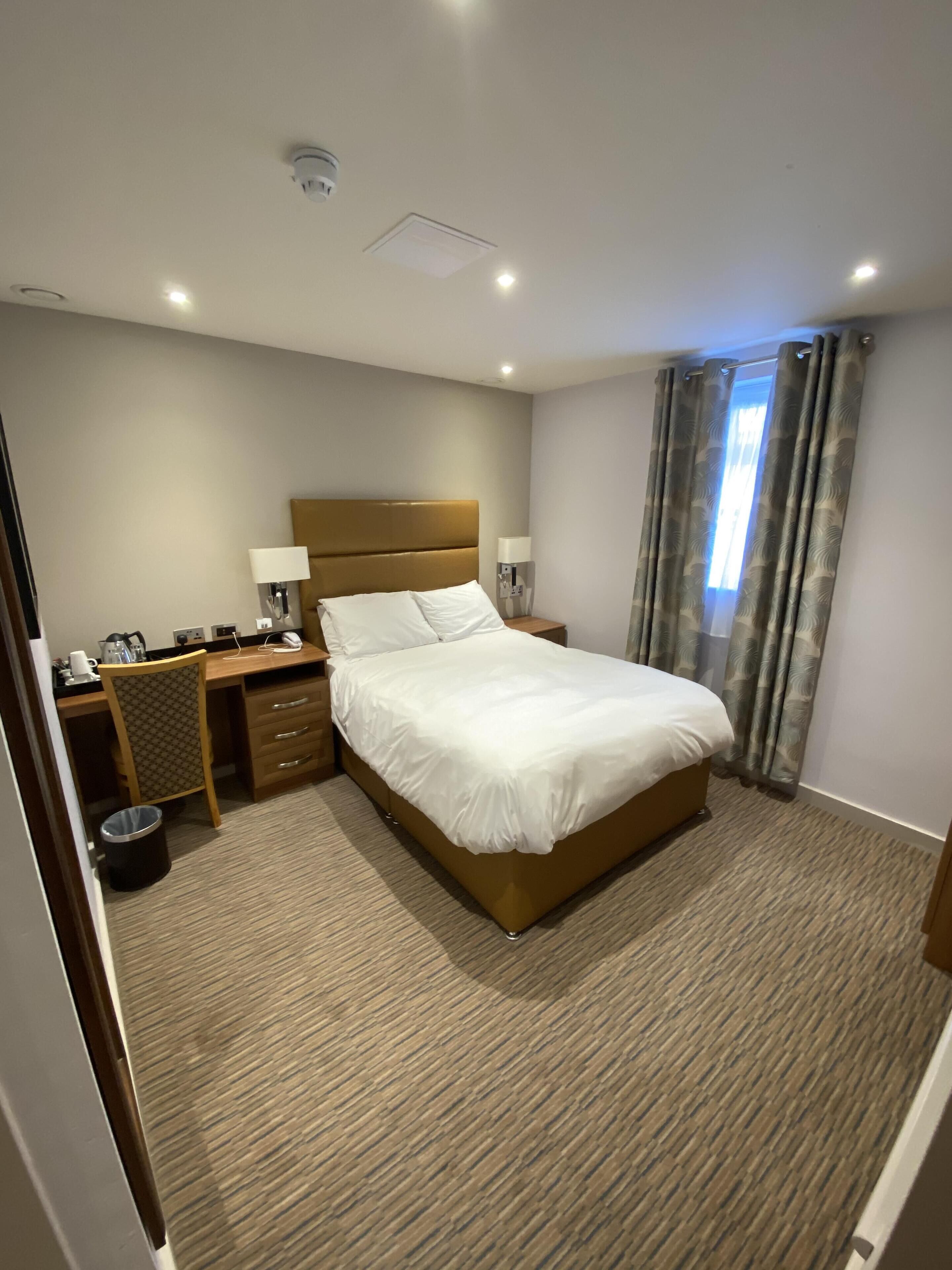 A hotel room with a large bed, a desk with a chair, a nightstand, a lamp, and a trash bin.