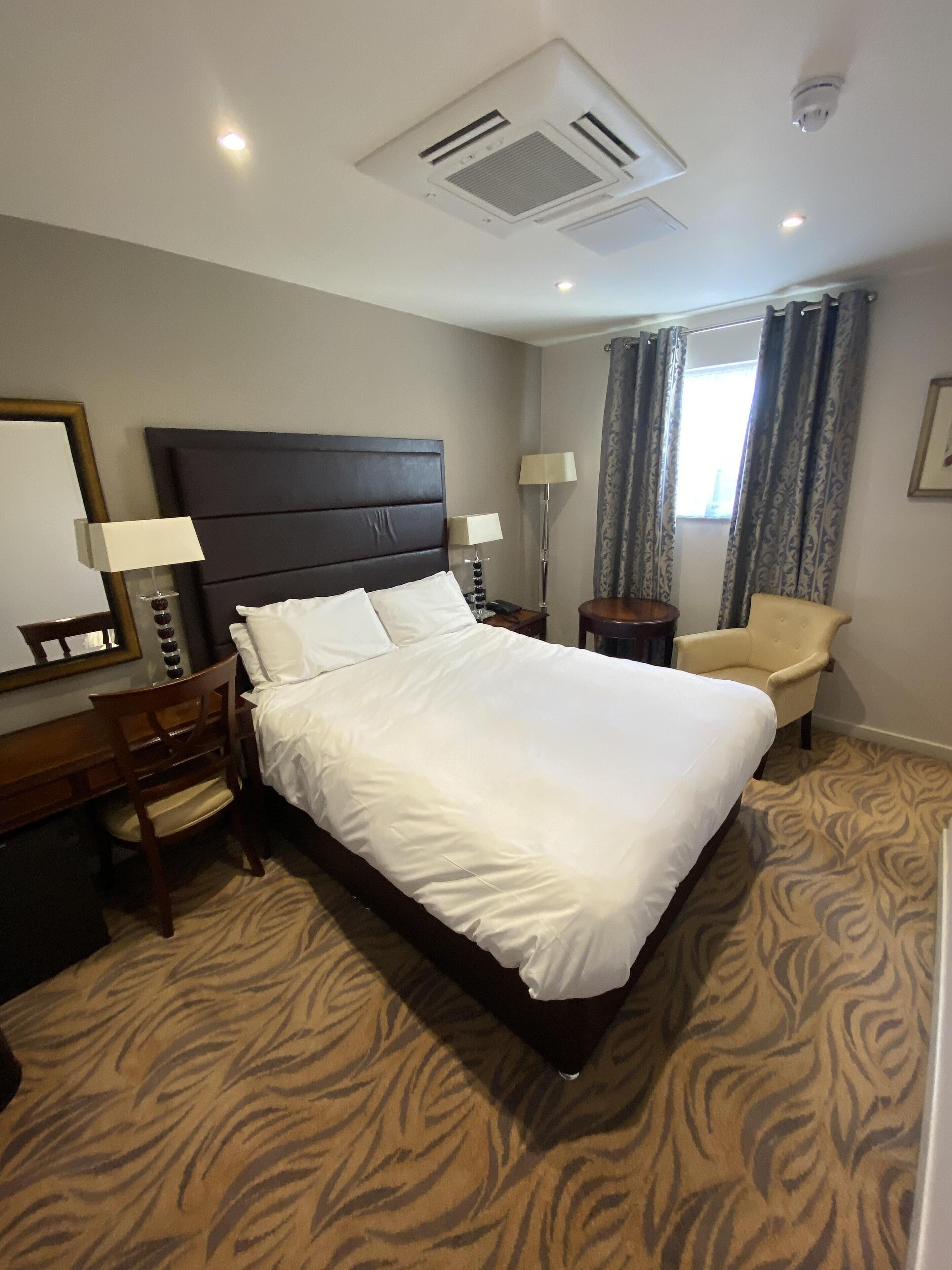 Une chambre d’hôtel avec un grand lit, un bureau, une chaise, une lampe et une fenêtre avec des rideaux.