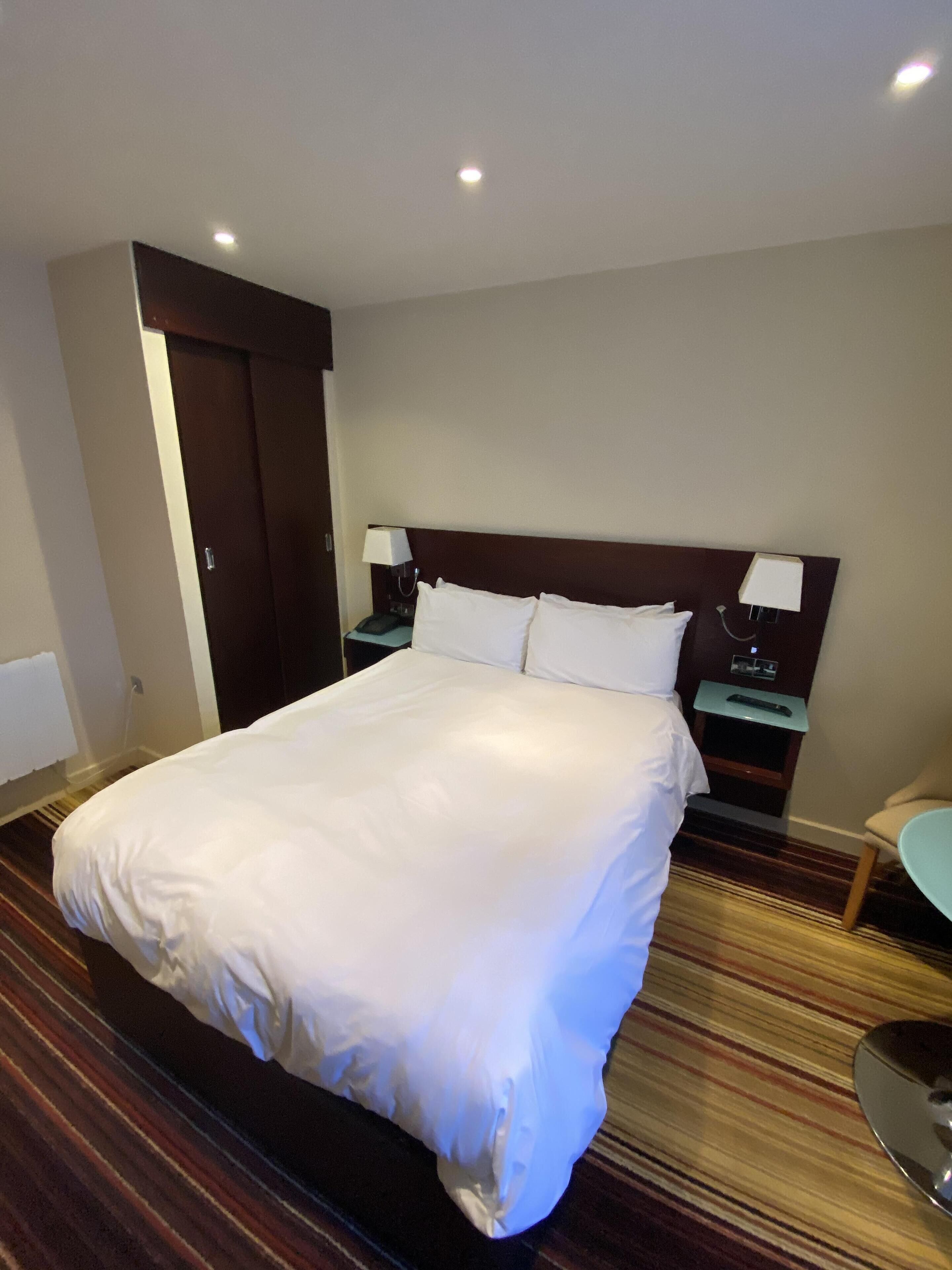 A hotel room with a large bed, two bedside tables with lamps, a wooden headboard, and a striped carpet.