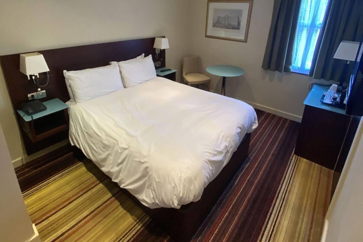 A hotel room with a large bed, bedside tables, a small round table, a chair, a desk, a lamp, a framed picture on the wall, and a window with curtains.