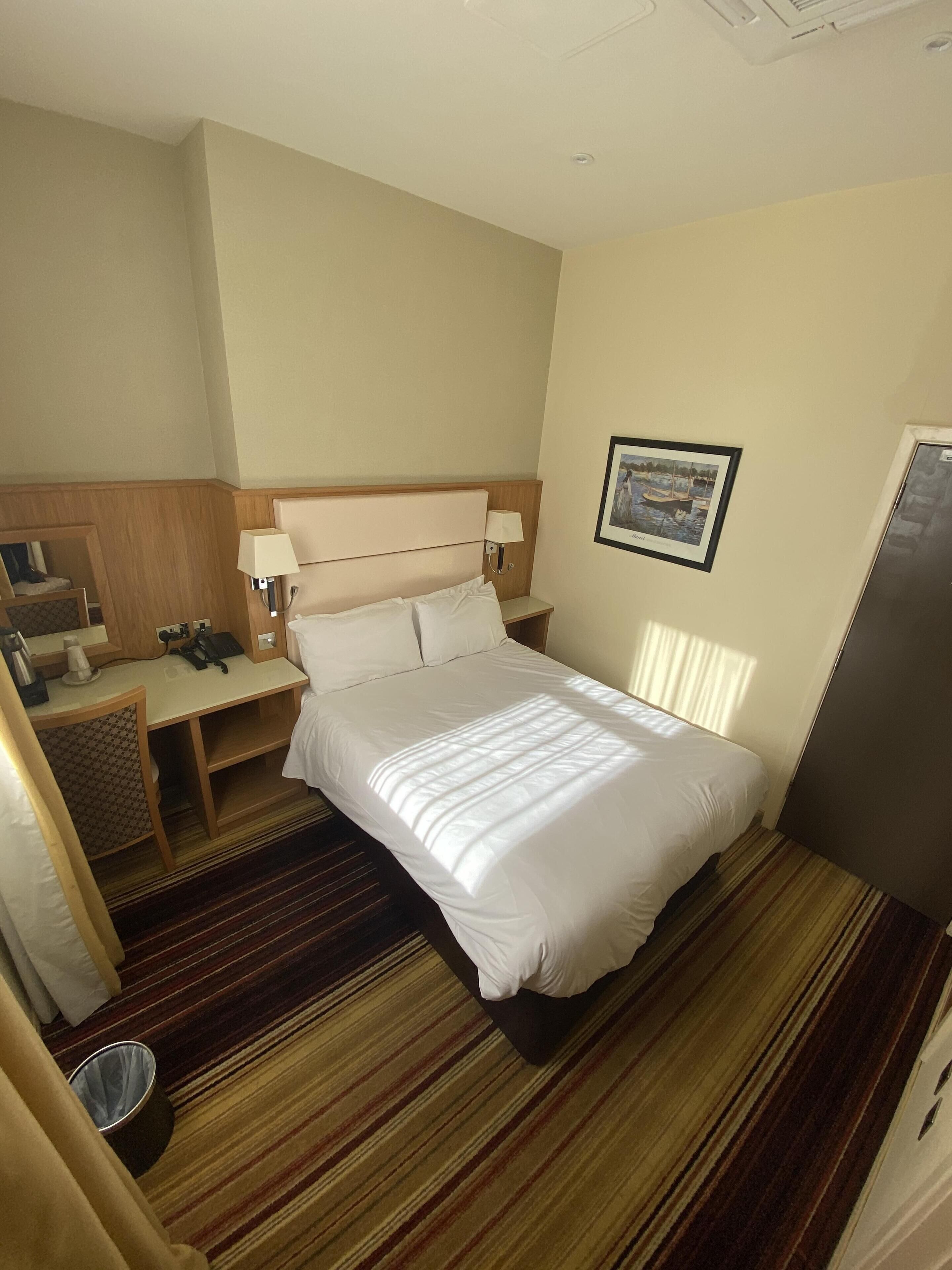 A hotel room with a bed, a desk with a lamp, a trash bin, and a framed picture on the wall.