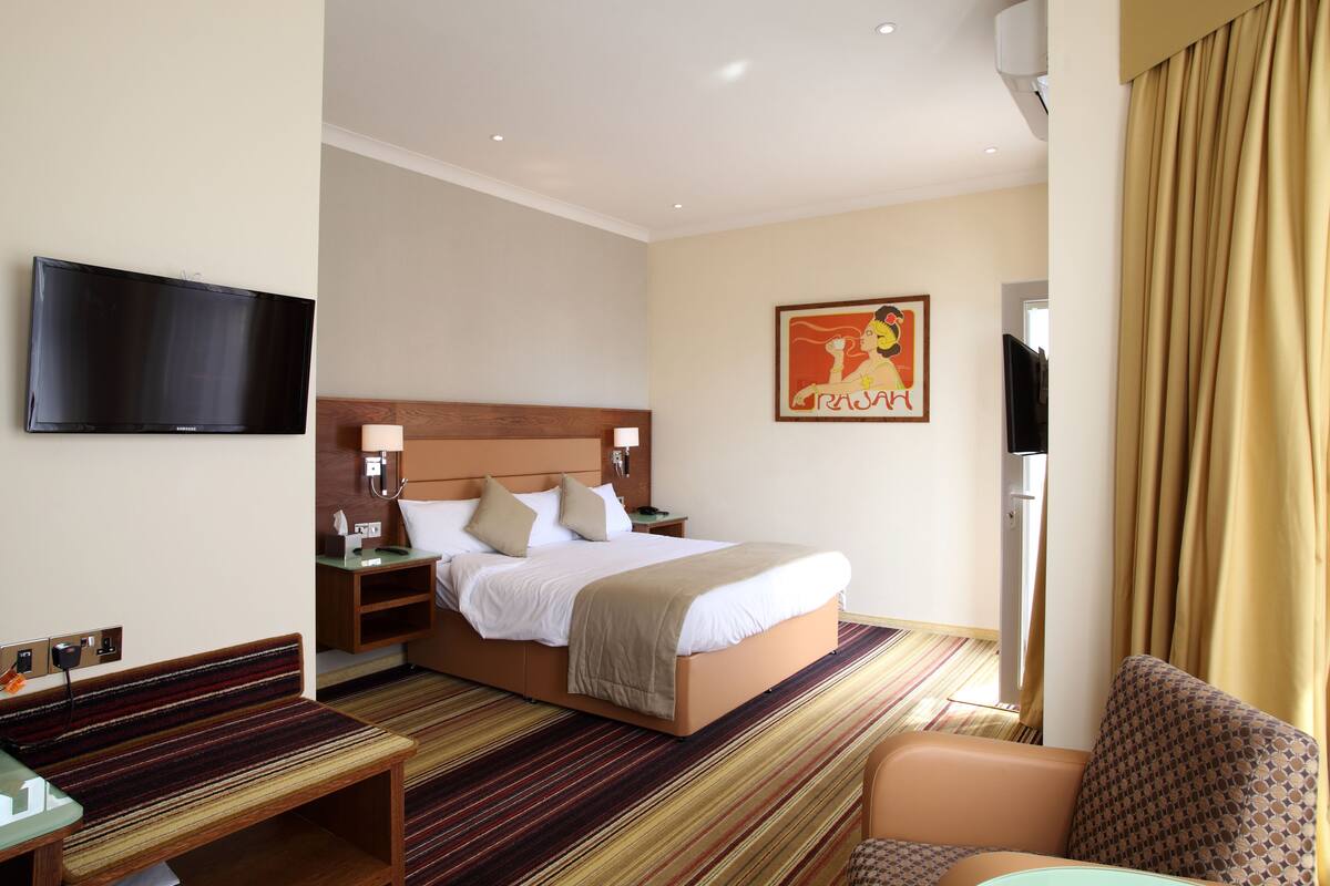 A hotel room with a bed, a TV, a bedside table, a chair, and a window with curtains.