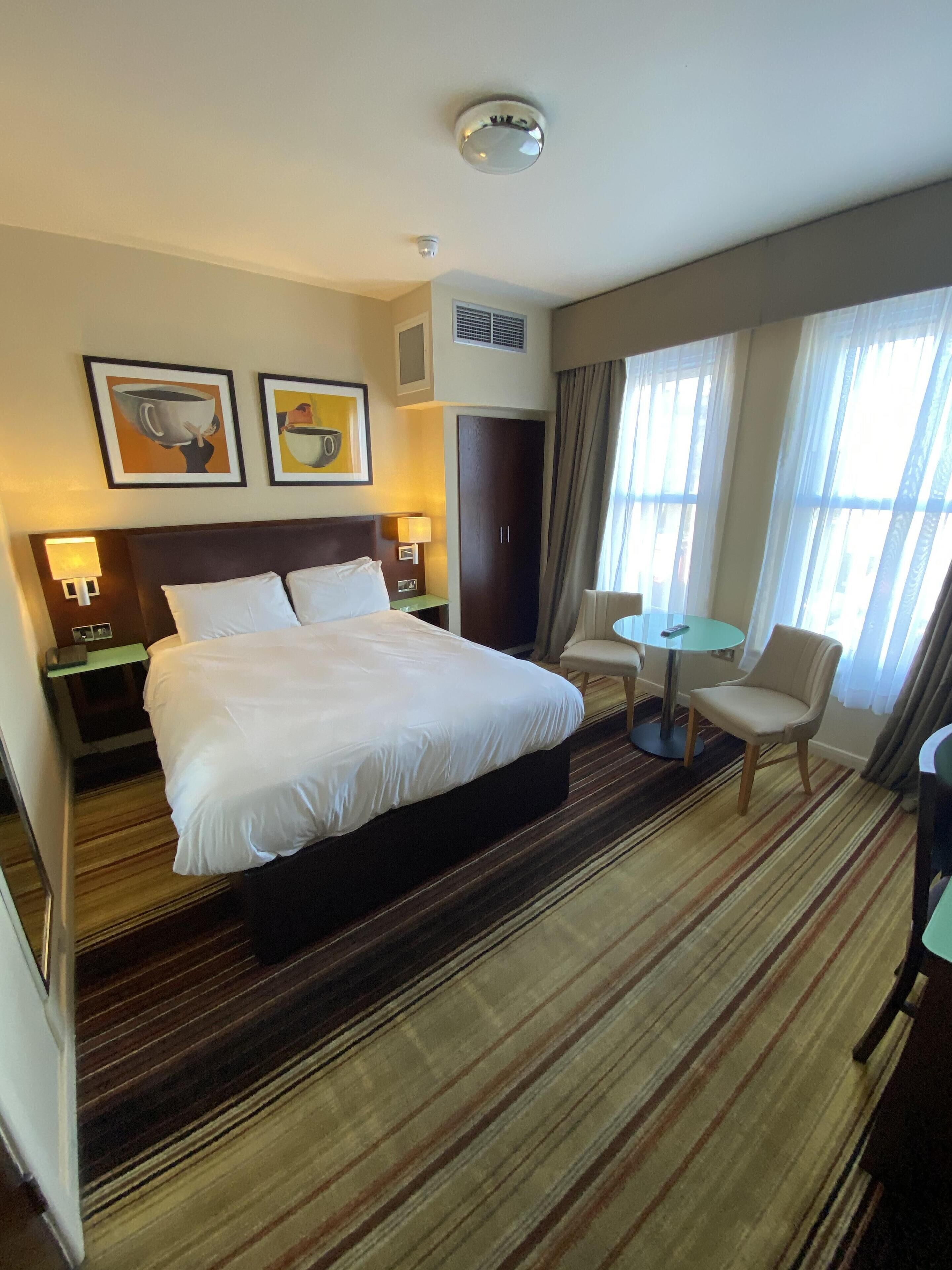 Hotelkamer met een groot bed, nachtkastje met lamp, een klein rond tafeltje met twee stoelen, en een kledingkast.