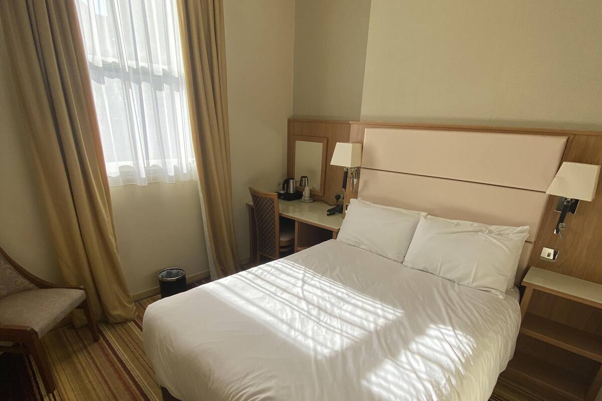 A hotel room with a large bed, a desk with a chair, a small table with a lamp, and a trash can.