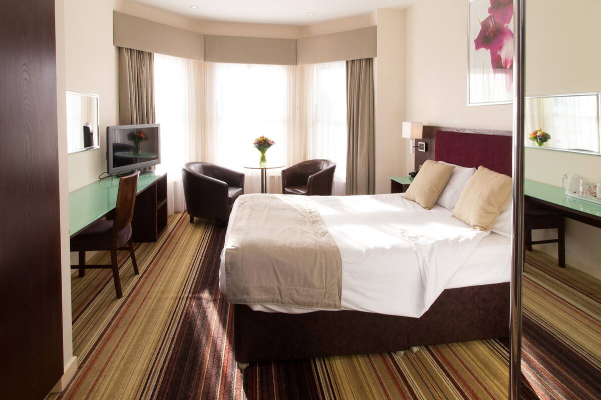 A hotel room with a large bed, a desk with a computer, a chair, and a small table with flowers.