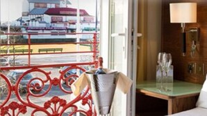 Hotellihuone, jossa on sänky, pieni pöytä lampun kanssa ja näkymä rakennukseen ikkunasta, jonka edessä on punainen kaide.