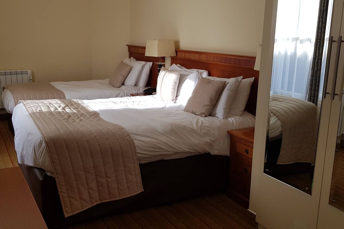 A hotel room with two beds, a wooden headboard, a bedside table with a lamp, a chair, and a view of the outside through a window.