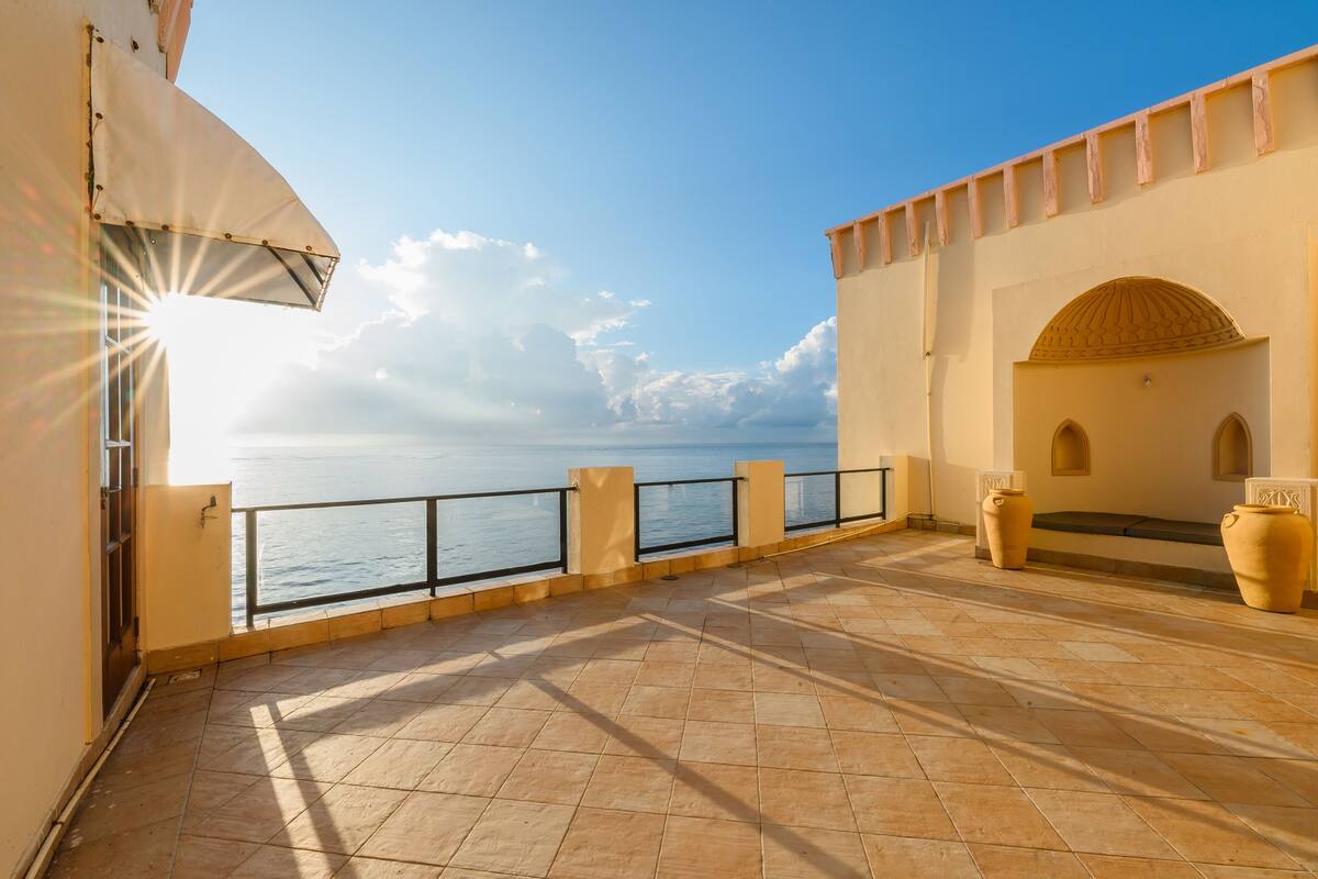 Une terrasse avec vue sur la mer, un sol carrelé et un bâtiment à la façade beige surmonté d’un grand parasol.