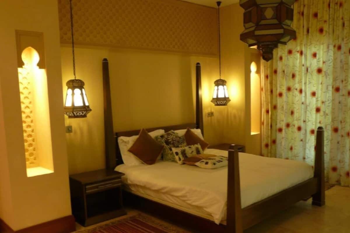 Une chambre dotée d’un lit à baldaquin, de lampes fixées au mur, d’un tapis à motifs et de rideaux ornés de motifs floraux.