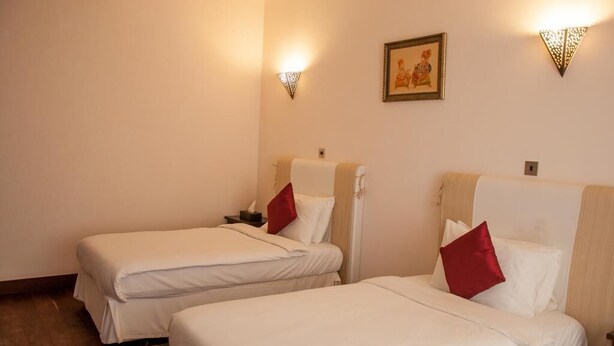 A hotel room with two single beds, each with a red pillow, a nightstand, and a wall-mounted light fixture.
