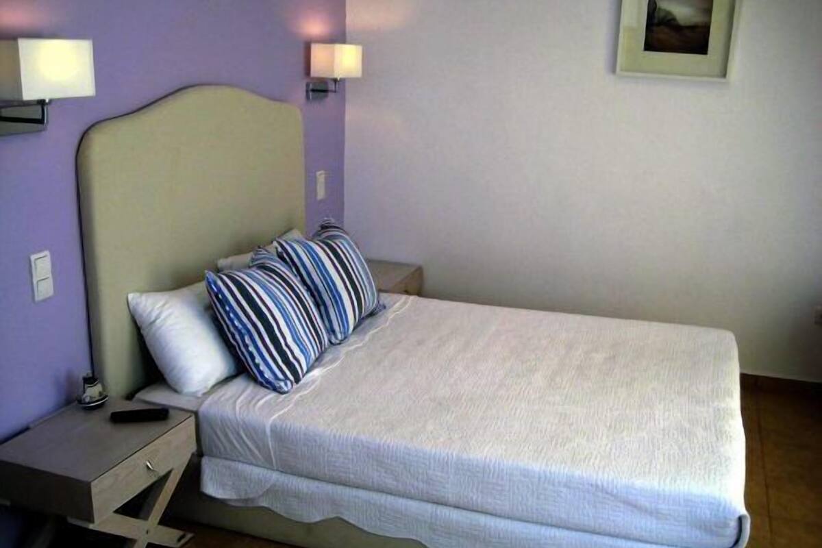 Un lit simple avec un matelas blanc et des oreillers à rayures, une table de chevet avec une lampe et un cadre photo fixé au mur.