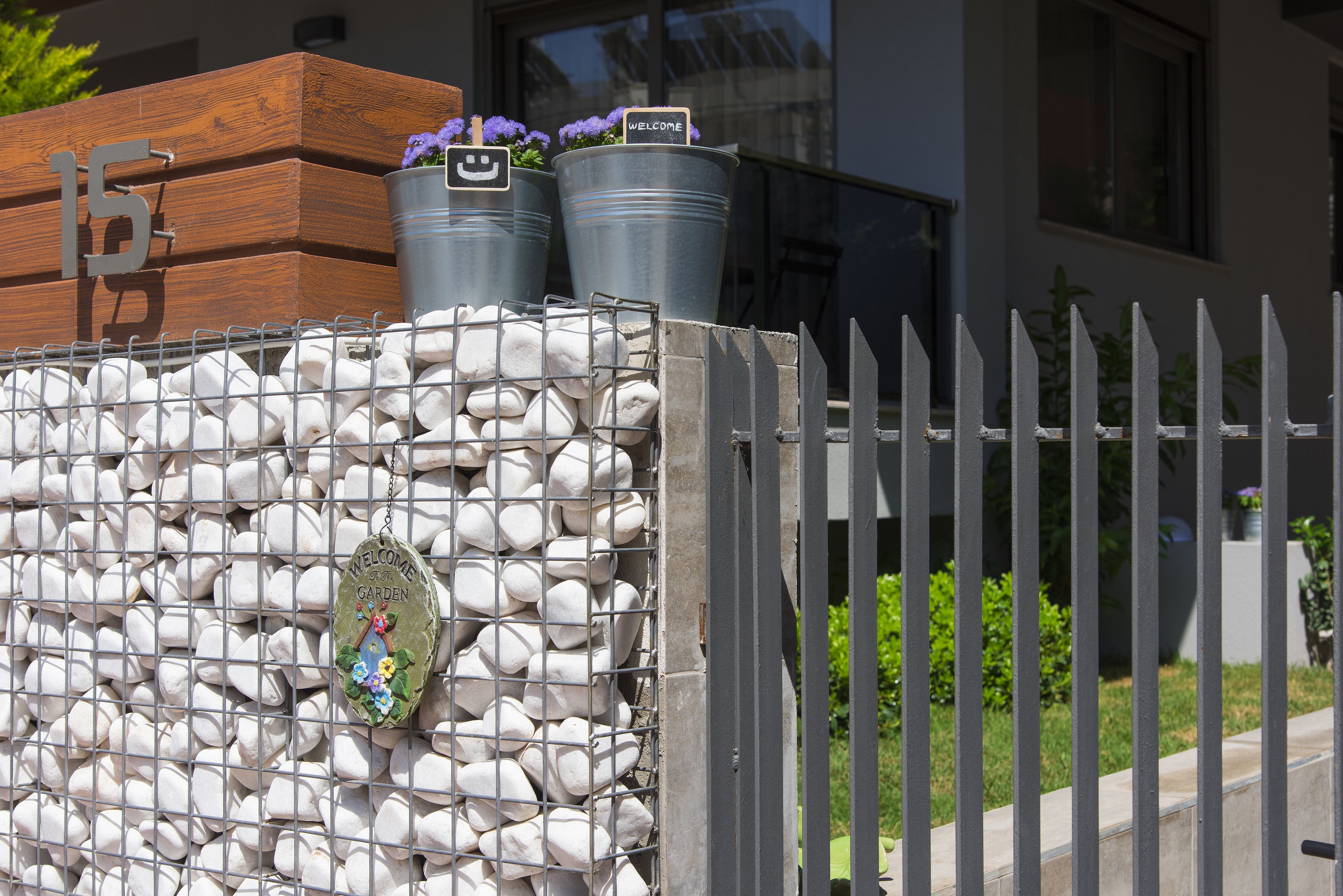 A stone wall with a metal gate, a wooden planter with flowers, and a welcome sign.