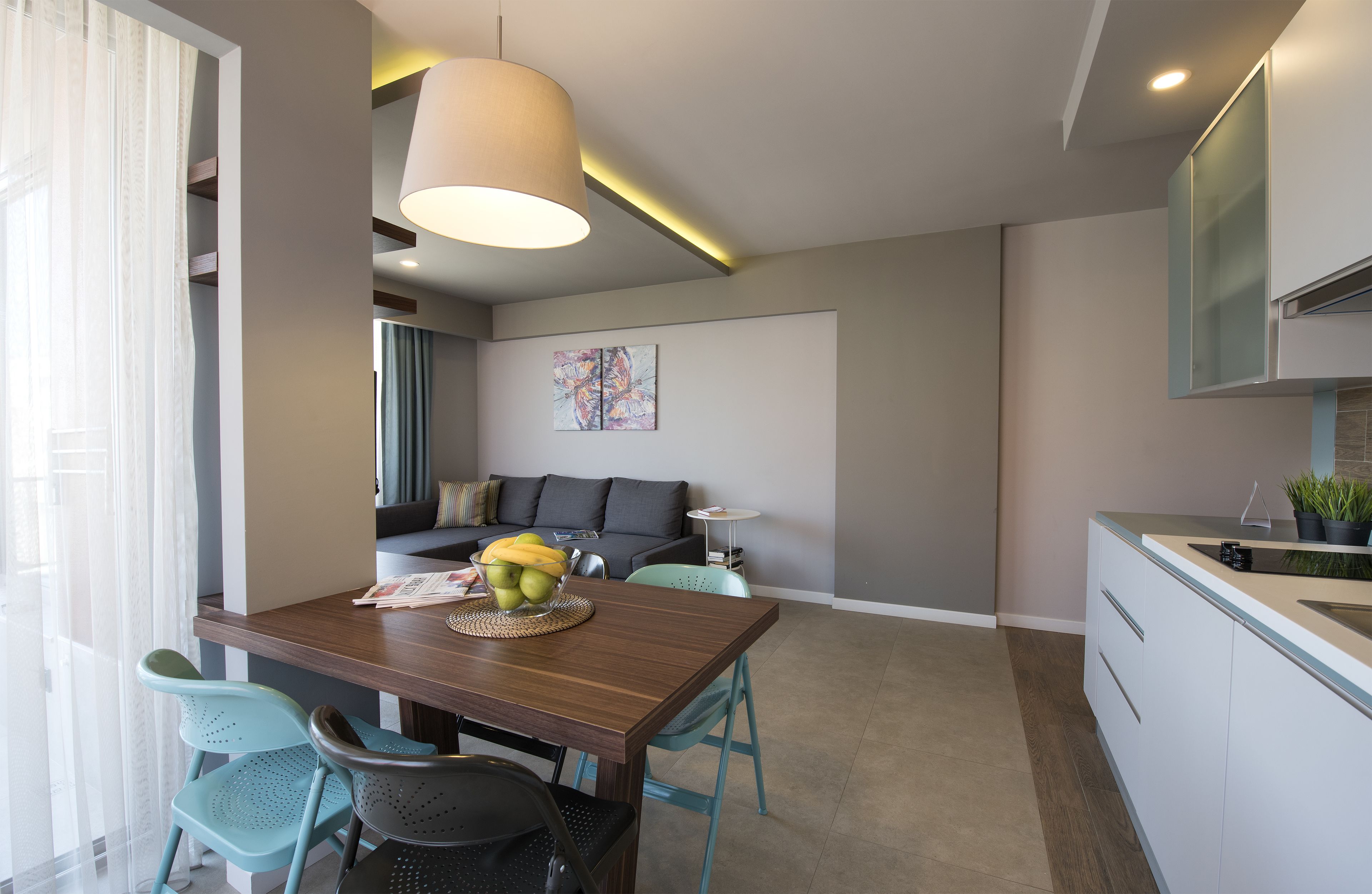 A modern kitchen with a sink, countertop, and cabinets. There is a dining area with a wooden table and chairs, and a sofa in the background.