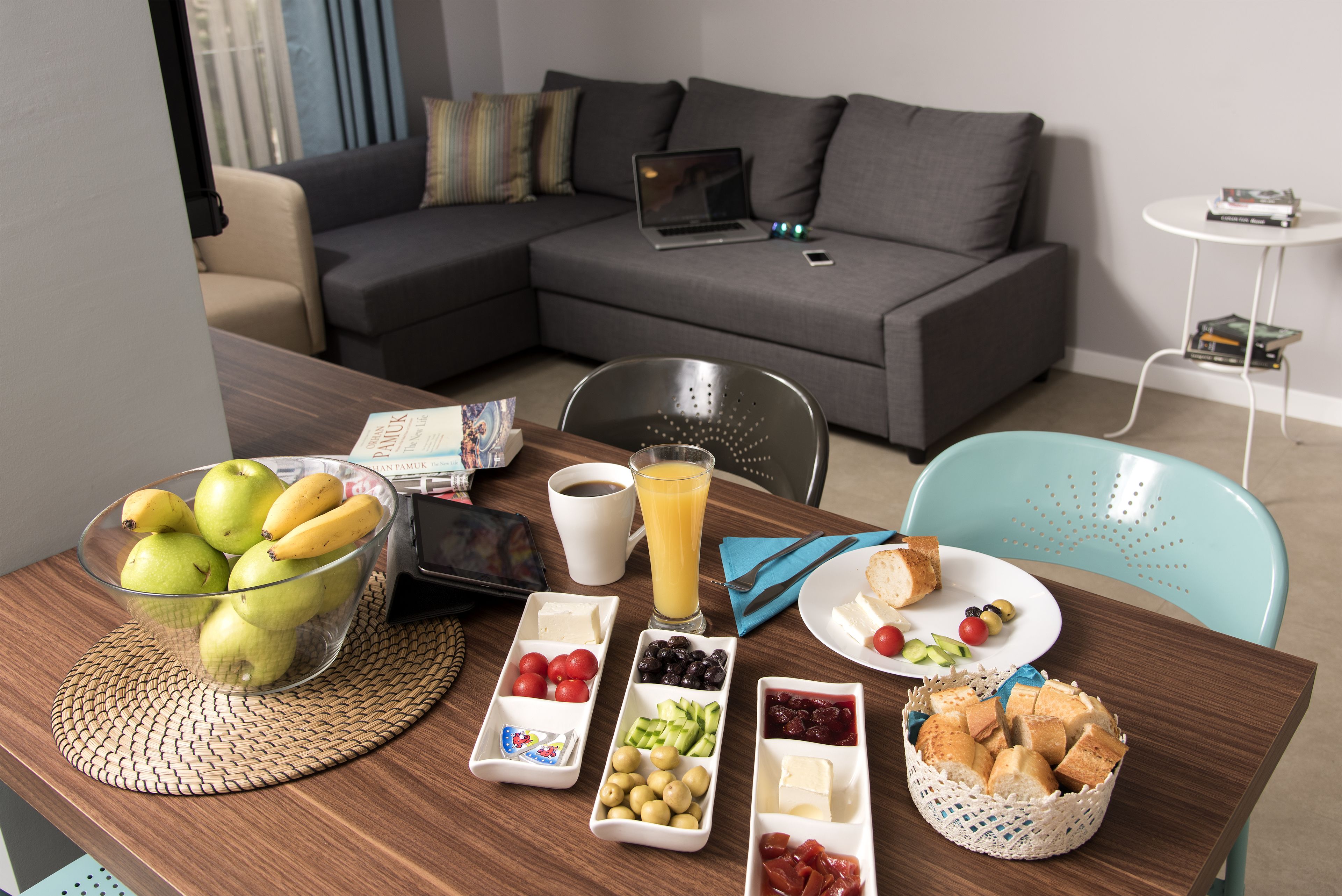 Desayuno con frutas, queso, pan y jugo sobre una mesa de madera en una sala de estar moderna con un sofá gris y una laptop encima.
