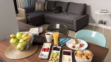 A breakfast setup with fruits, cheese, bread, and juice on a wooden table in a modern living room with a grey sofa and a laptop on it.