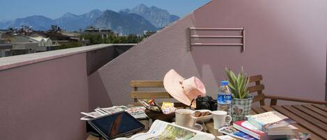 A balcony with a view of mountains, a wooden bench, a table with books, a camera, a water bottle, a hat, and a potted plant.