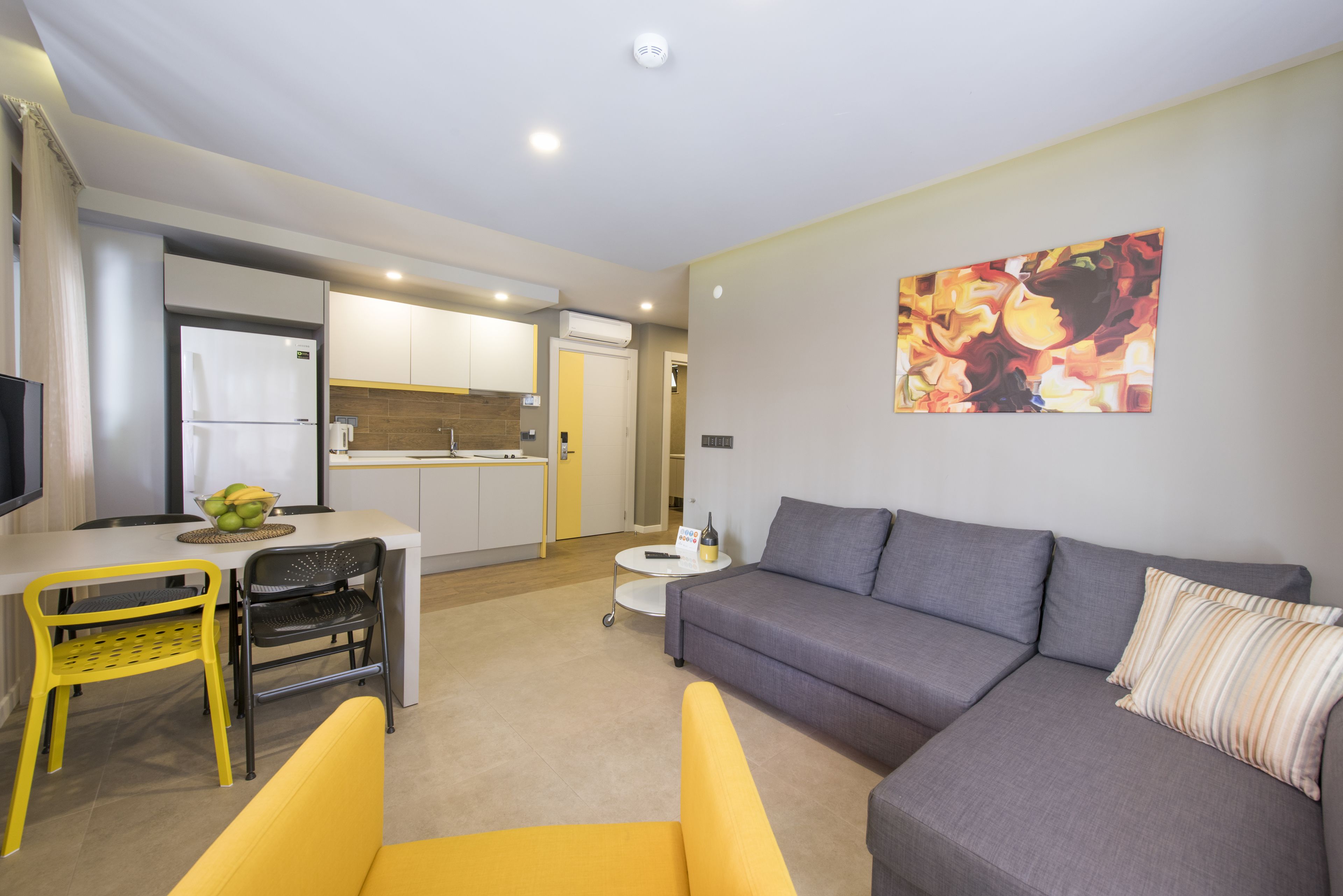Una sala de estar moderna con un sofá gris, sillas amarillas, una mesa de comedor con un cuenco de fruta y una cocina al fondo.