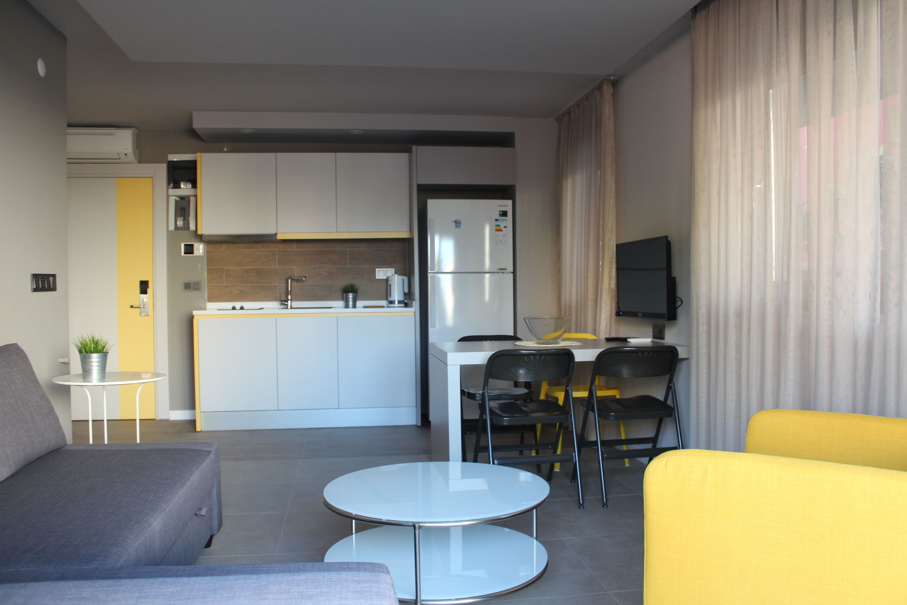 A modern living room with a grey sofa, a yellow armchair, and a small round table. A kitchen with white cabinets and a refrigerator is visible in the background.