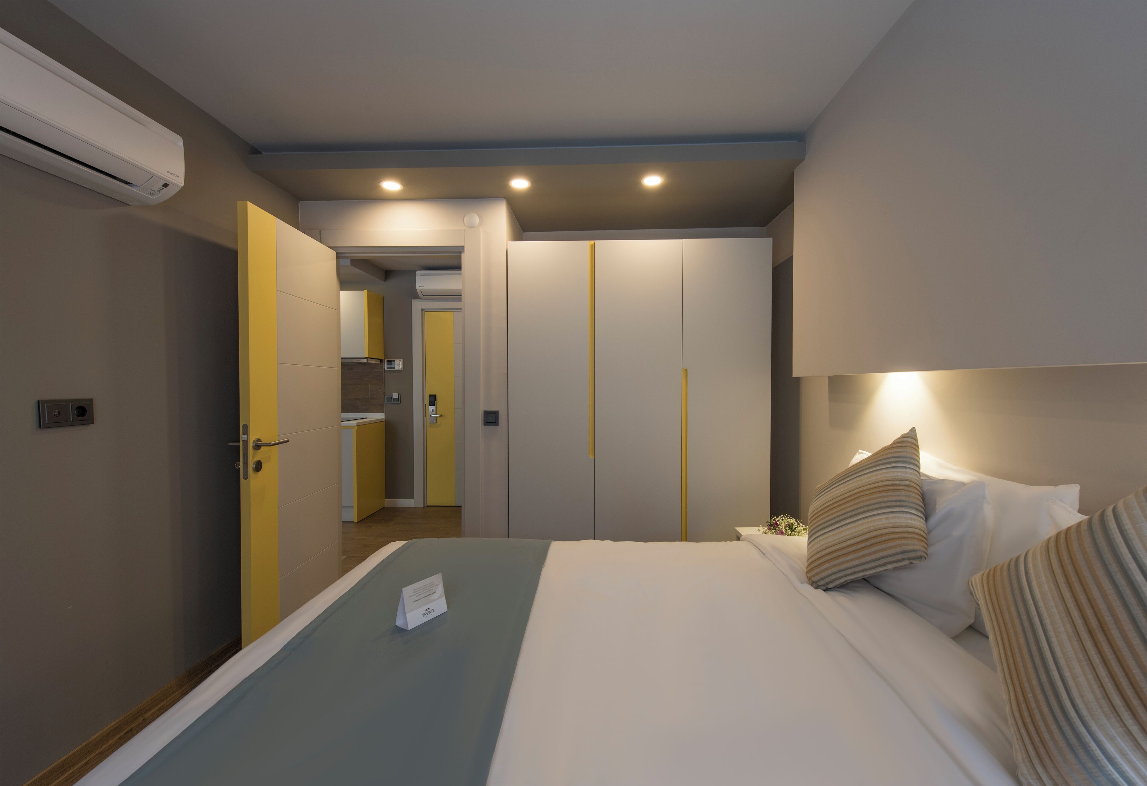 Habitación de hotel con cama, mesita de noche y una pequeña mesa auxiliar. Hay una puerta que da a un baño y una kitchenette.
