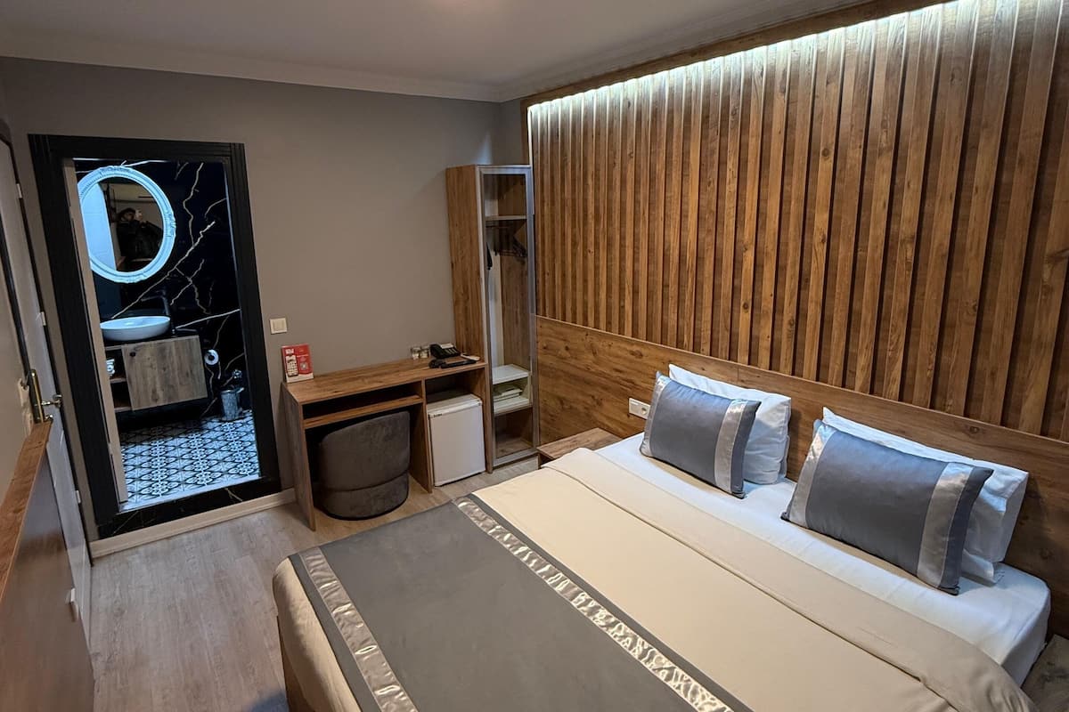 A modern hotel room with a large bed, wooden headboard, a built-in wardrobe, a vanity with a mirror, and a small desk with a chair.