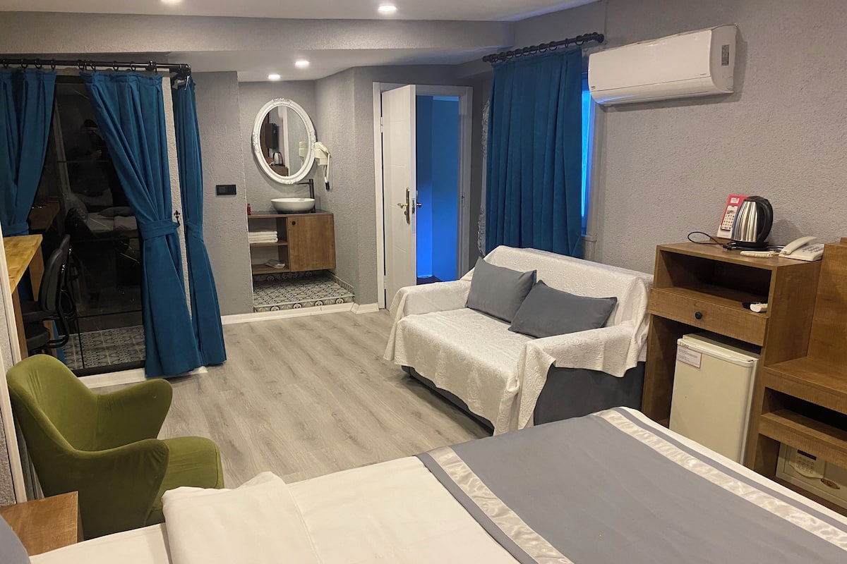 A hotel room with a bed, a green chair, a desk with a computer, a small bathroom with a sink and mirror, and a wardrobe.