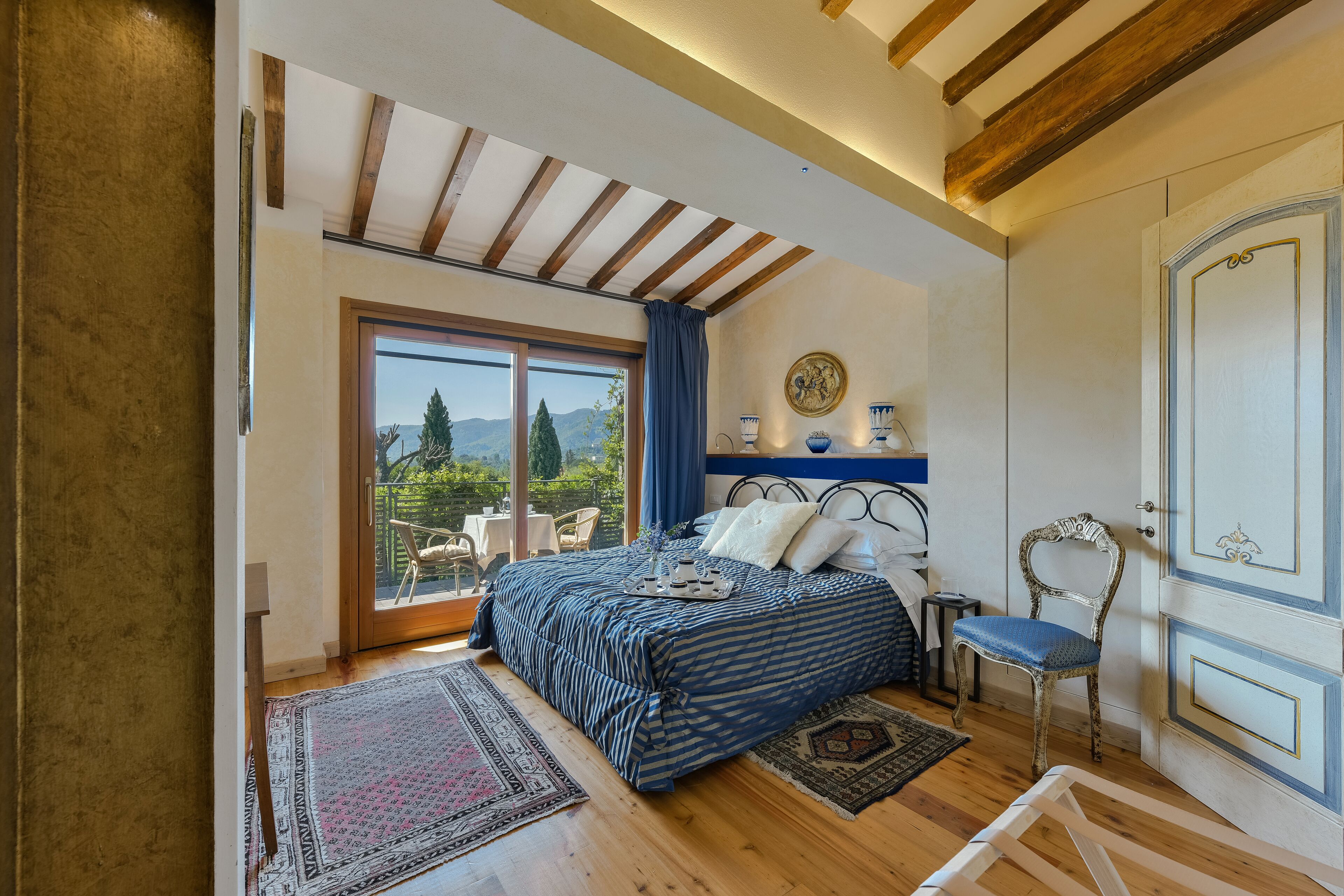 Un dormitorio con un ventanal grande, una cama con colcha azul y blanca a rayas, techo de madera y vistas al exterior.