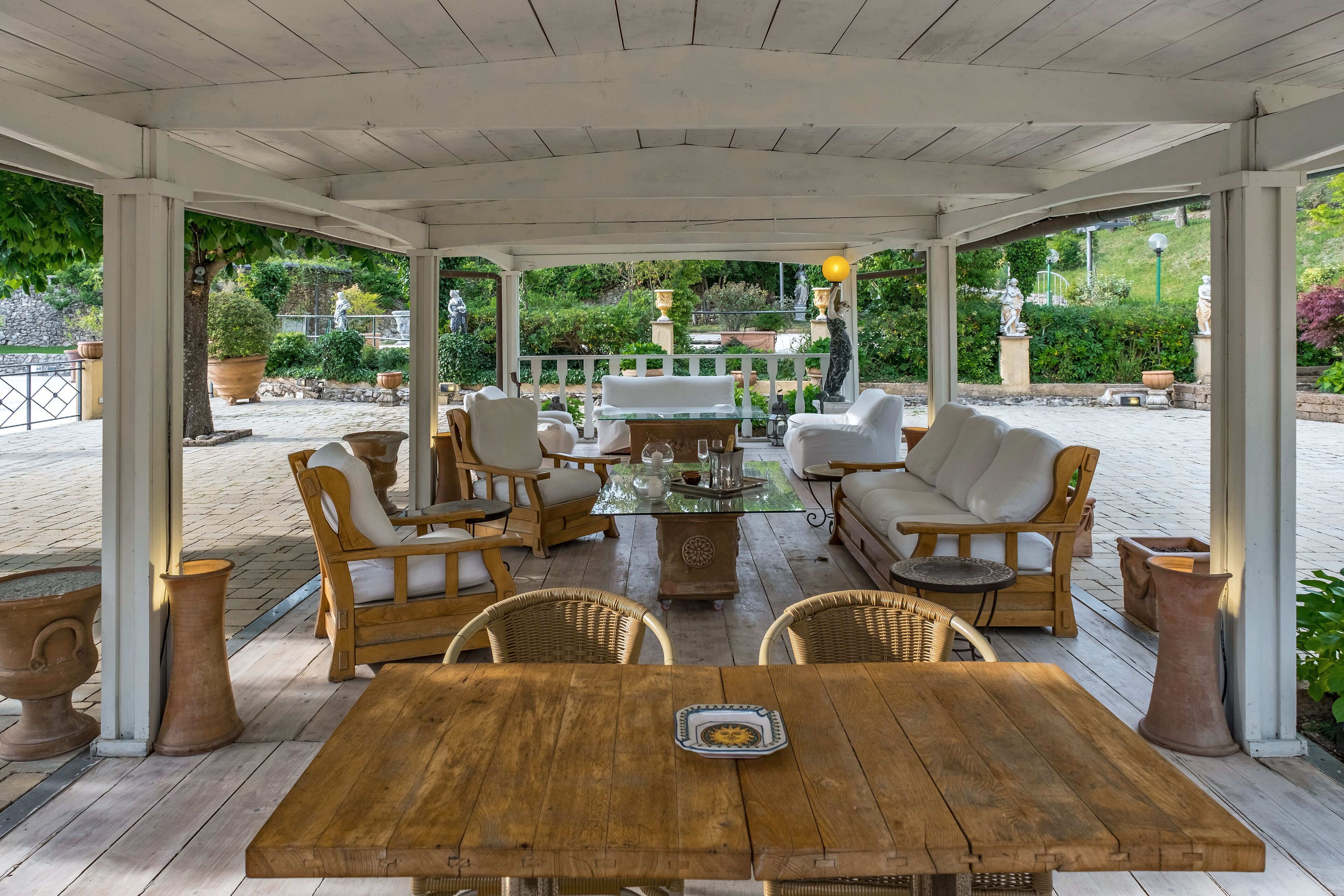 Un patio techado al aire libre con mobiliario de madera, una mesa de vidrio y macetas de terracota.