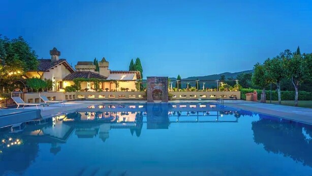 Una amplia piscina al aire libre con el reflejo de un edificio y árboles, rodeada de zonas de descanso bien iluminadas y un cielo nocturno despejado.
