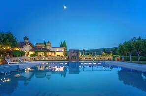 Une grande piscine extérieure où se reflètent un bâtiment et des arbres, entourée d’espaces de détente bien éclairés et sous un ciel nocturne dégagé.