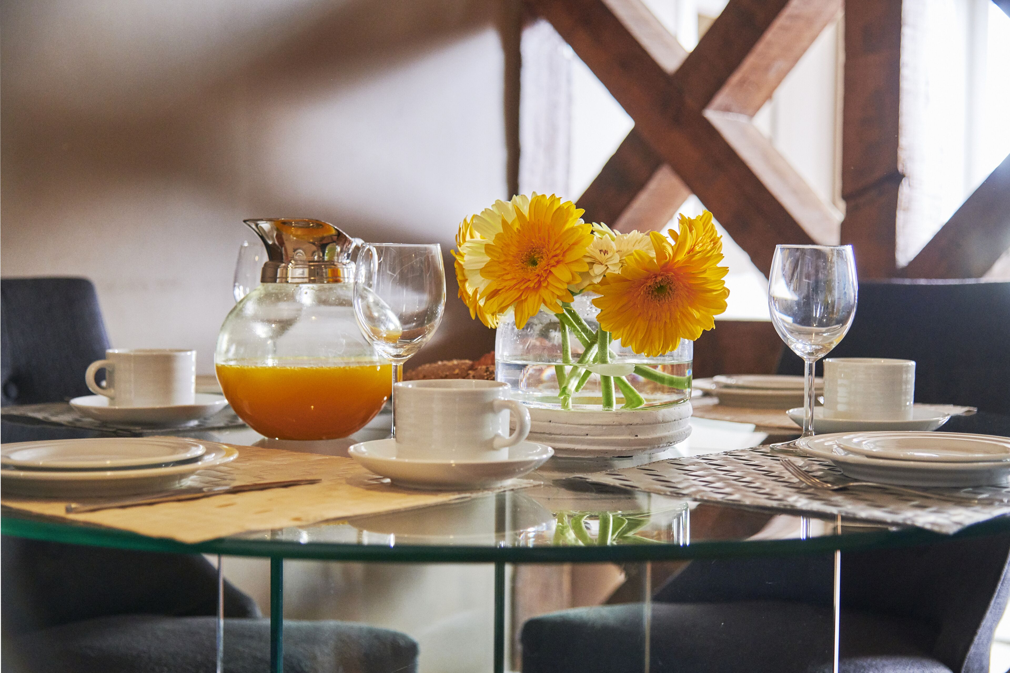 A breakfast table set with plates, cups, a pitcher of orange juice, a vase with yellow flowers, and a wine glass.