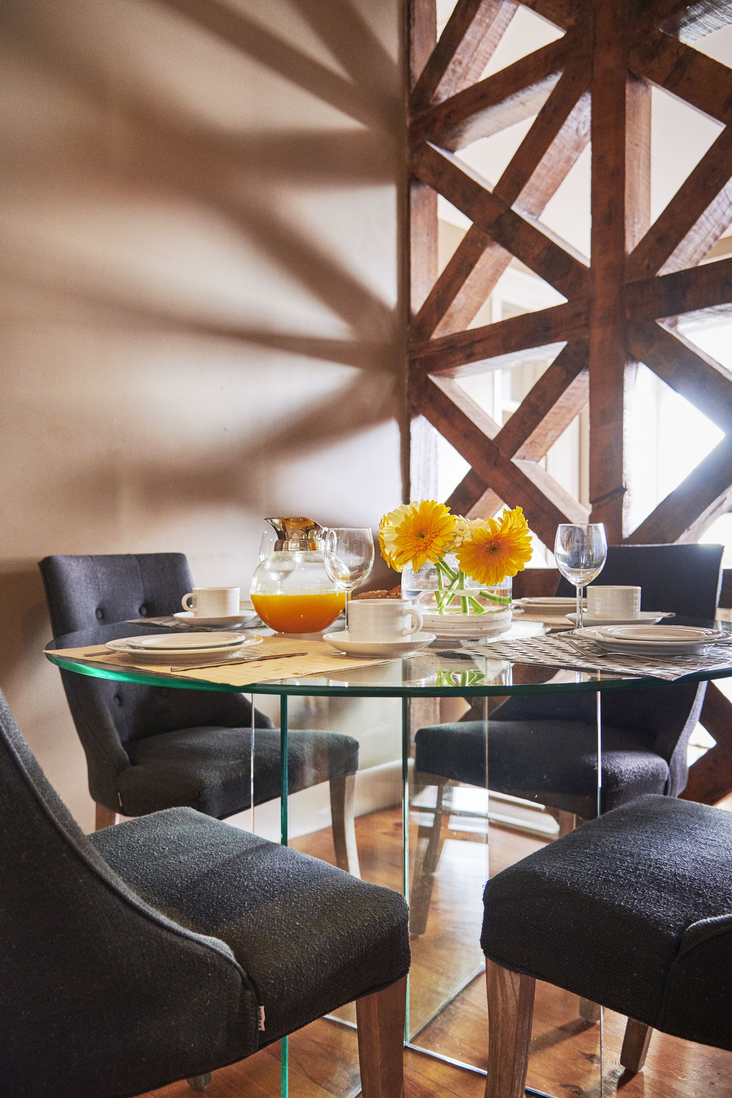 Een eettafel met een glazen blad, omringd door donker beklede stoelen. Op de tafel staat een karaf sinaasappelsap, een glas water en een vaas met gele bloemen.