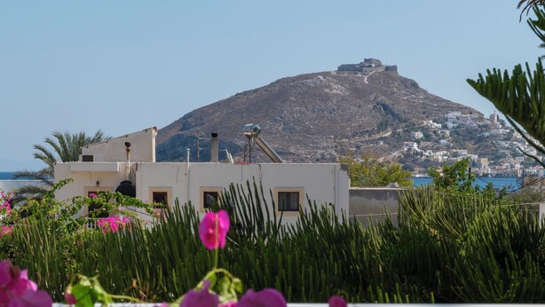 A table with a bottle, glass, and hat, surrounded by greenery and a view of a hill with buildings. - Irene Hotel (Leros)