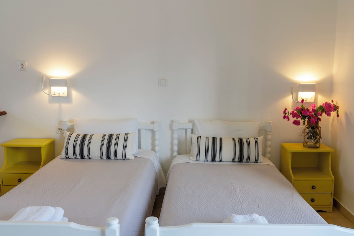 Una habitación de hotel con dos camas, cada una con cabecero blanco y almohada a rayas. Hay mesitas de noche con cajones amarillos y un florero con flores rosas sobre la mesa de la derecha.