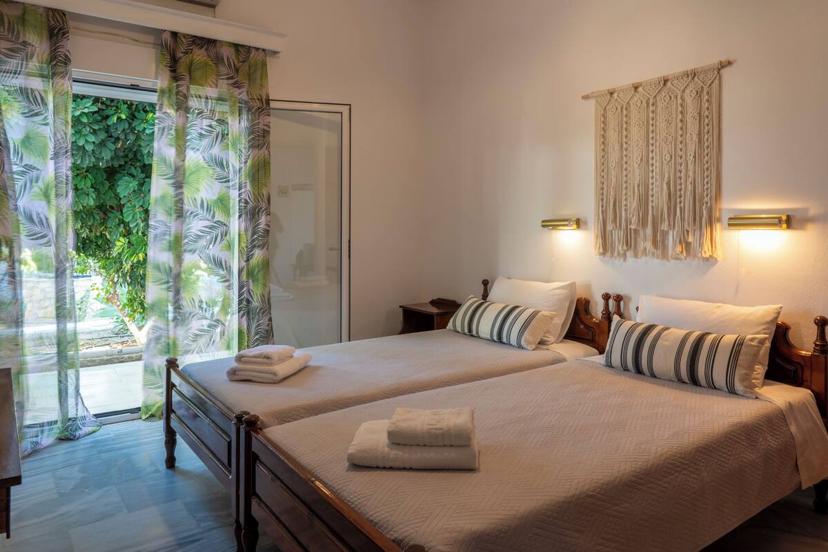 Una cama doble con estructura de madera, dos mesitas de noche, un colgador de macramé en la pared y vistas a la vegetación a través de una puerta de cristal.