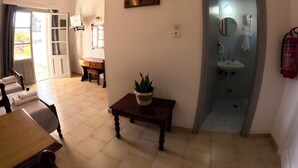 A room with a dining table, chairs, a small wooden table with a plant, a mirror, and a bathroom with a sink and towel rack. - Irene Hotel (Leros)