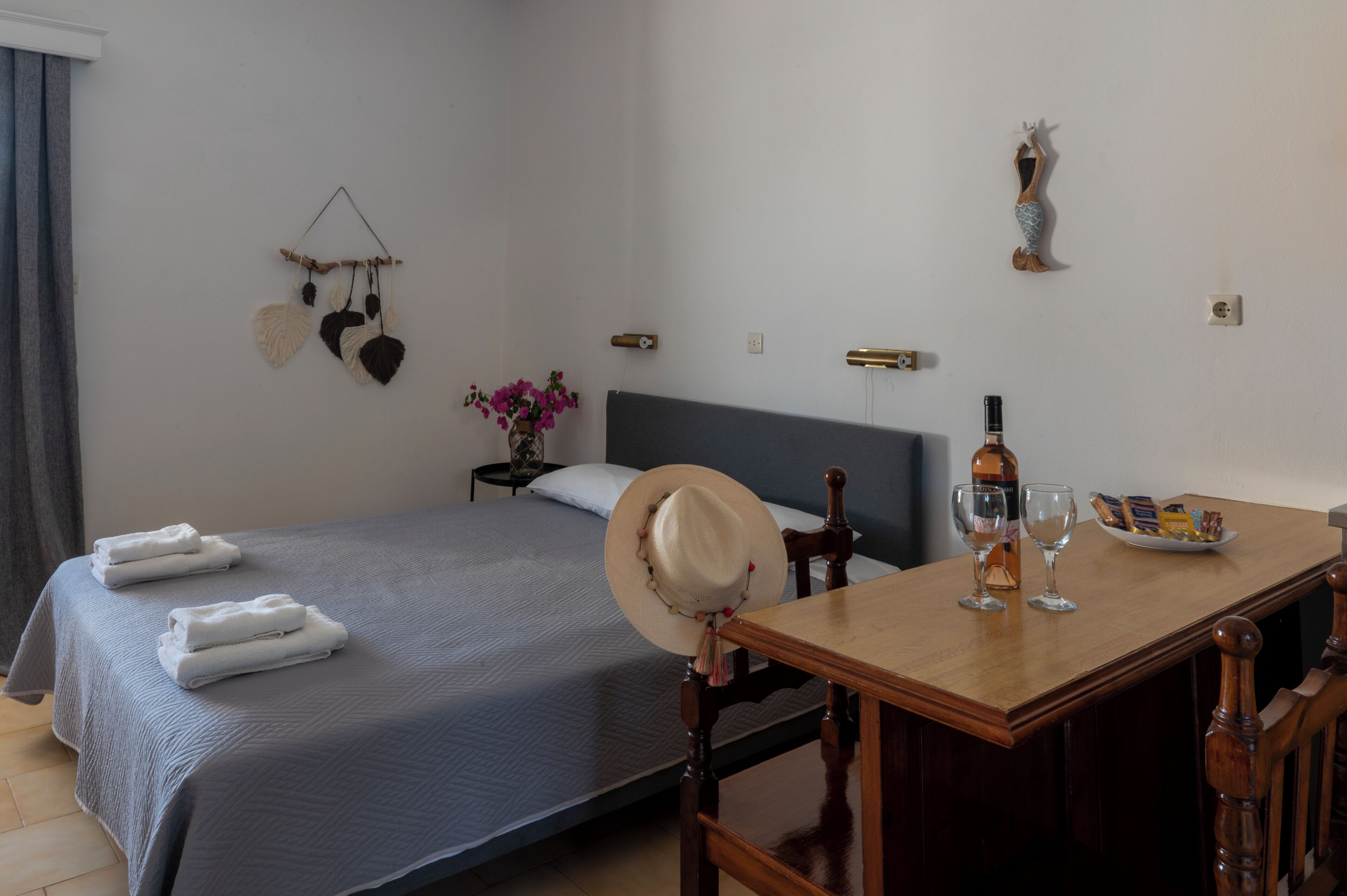 Een net opgemaakt bed met opgevouwen handdoeken, een houten tafel met glazen en een fles, en een decoratief object aan de muur.